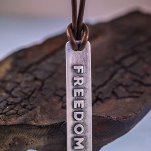 May include: A silver pendant necklace with the word "FREEDOM" engraved vertically. The pendant hangs from a brown leather cord. The background is a dark, textured surface.