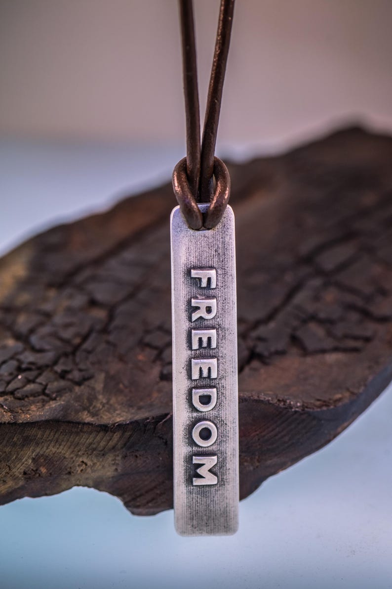 May include: A silver pendant necklace with the word "FREEDOM" engraved vertically. The pendant hangs from a brown leather cord. The background is a dark, textured surface.