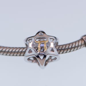 K&ouml;nnte beinhalten: Ein silbernes Armband mit einem silbernen und goldenen Anh&auml;nger in Form eines Davidsterns. Der Anh&auml;nger hat einen goldenen hebr&auml;ischen Buchstaben in der Mitte.