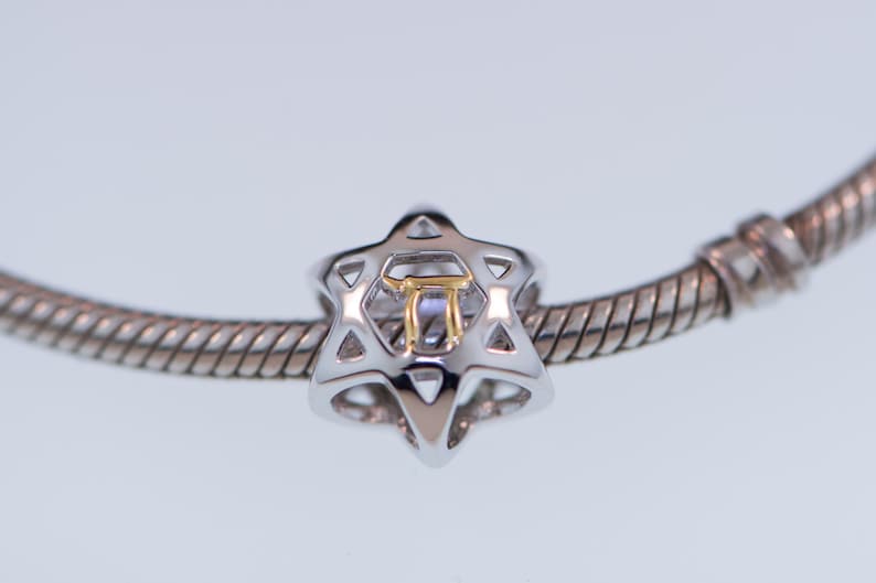 K&ouml;nnte beinhalten: Ein silbernes Armband mit einem silbernen und goldenen Anh&auml;nger in Form eines Davidsterns. Der Anh&auml;nger hat einen goldenen hebr&auml;ischen Buchstaben in der Mitte.