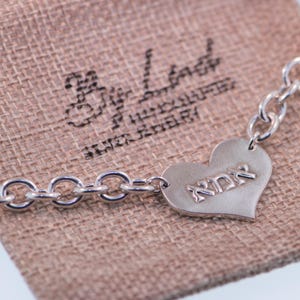 K&ouml;nnte beinhalten: Ein silbernes Kettenarmband mit herzf&ouml;rmigem Anh&auml;nger. Der Anh&auml;nger tr&auml;gt das Wort "אמא" eingraviert. Das Armband liegt auf einem beigen Stoffbeutel.
