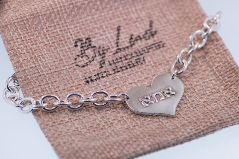 K&ouml;nnte beinhalten: Ein silbernes Kettenarmband mit herzf&ouml;rmigem Anh&auml;nger. Der Anh&auml;nger tr&auml;gt das Wort "אמא" eingraviert. Das Armband liegt auf einem beigen Stoffbeutel.