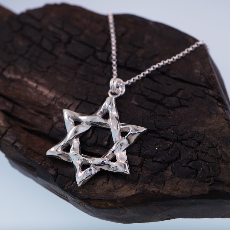 Puede incluir: Un collar con un colgante de estrella de David de plata con un dise&ntilde;o trenzado. El collar est&aacute; sobre una superficie de madera marr&oacute;n oscura.