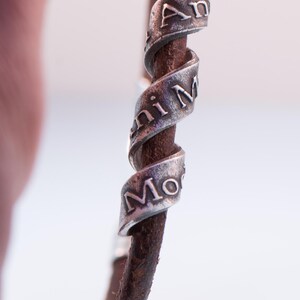May include: A brown leather bracelet with three silver bands spiralling around it. The silver bands have the engraved words "Ani Mi Mo". The bracelet is held between two fingers.