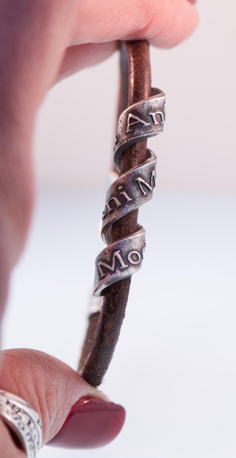 May include: A brown leather bracelet with three silver bands spiralling around it. The silver bands have the engraved words "Ani Mi Mo". The bracelet is held between two fingers.