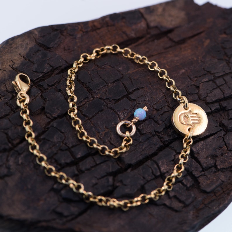 May include: A gold chain bracelet with a hamsa hand charm and a light blue opal bead. The bracelet is on a dark brown wooden surface.