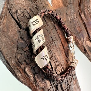 May include: A brown leather braided bracelet with silver charms. The charms feature Hebrew lettering and a Star of David.