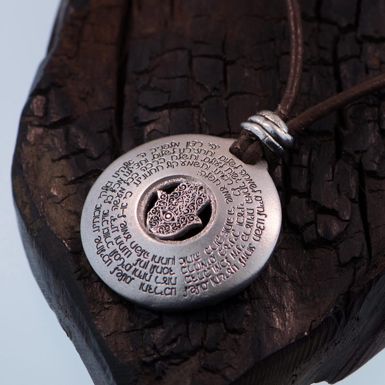 May include: A silver-colored pendant necklace with a circular design and a central Hamsa hand symbol. The pendant has engraved text around the edge and hangs from a brown leather cord.