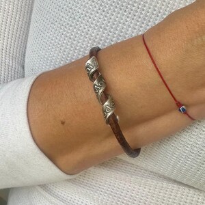 May include: A brown leather bracelet with three silver spiral accents, each engraved with a word. A thin red string bracelet with a small blue bead is also visible.