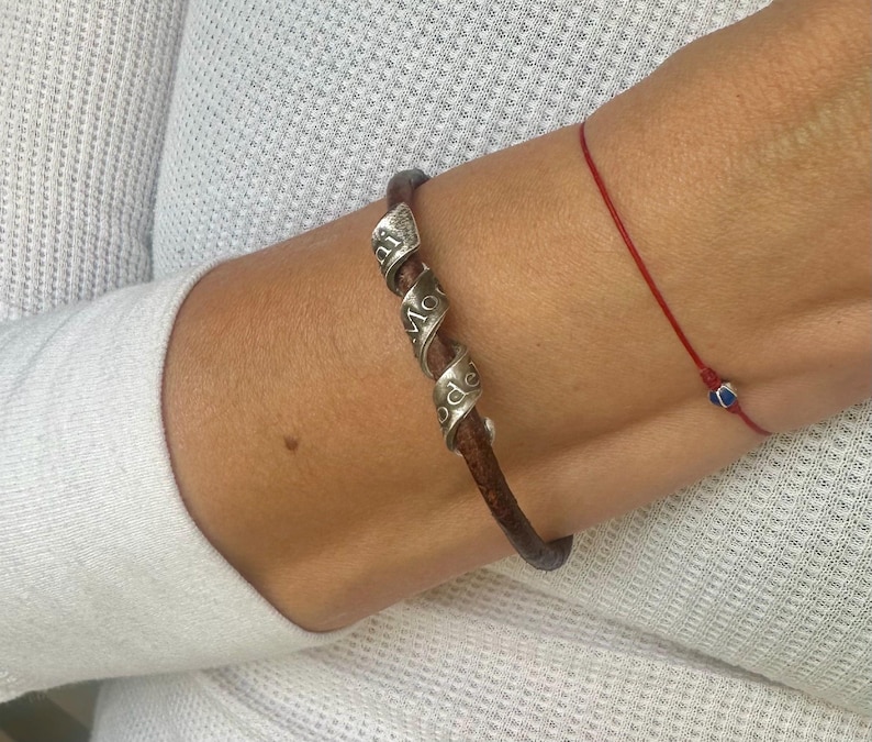 May include: A brown leather bracelet with three silver spiral accents, each engraved with a word. A thin red string bracelet with a small blue bead is also visible.