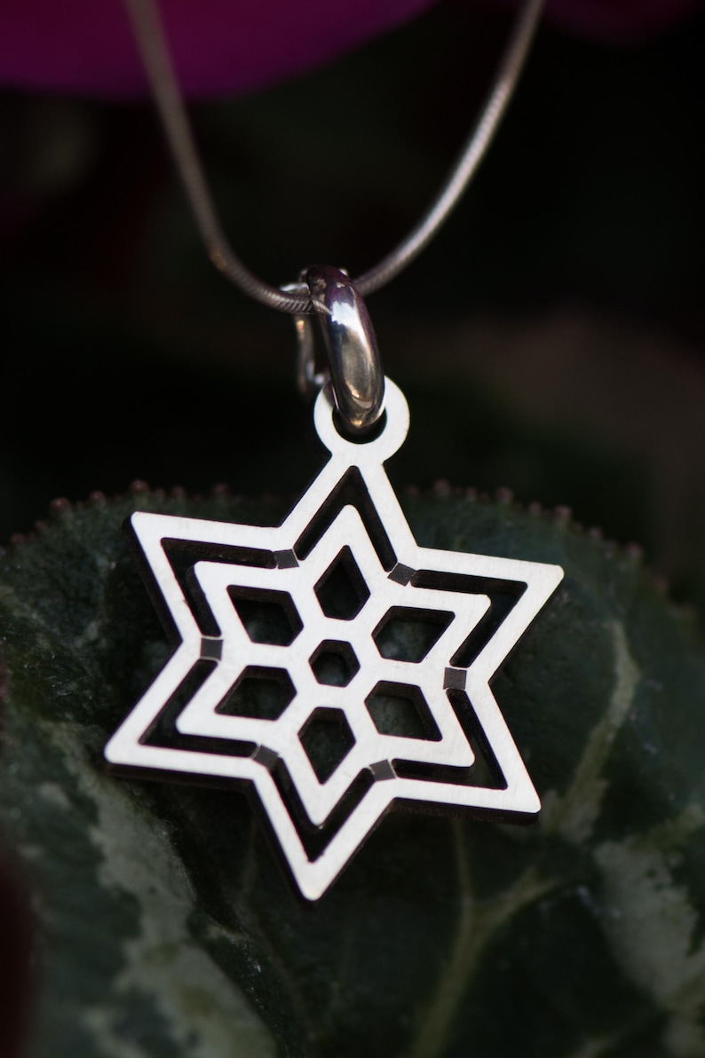 May include: A silver-colored pendant necklace featuring a six-pointed star design. The star has a geometric pattern with a central hexagon and surrounding diamond shapes. The pendant hangs from a thin, silver chain.