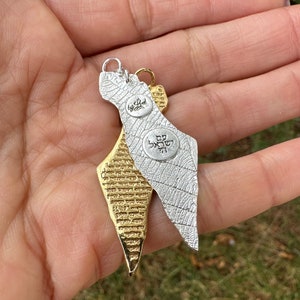 May include: Two pendant necklaces in the shape of Israel. One is silver-colored with a leaf-like texture and the other is gold-colored with a textured surface. Both have small, round charms with Hebrew text. The pendants are hanging from a person's hand.