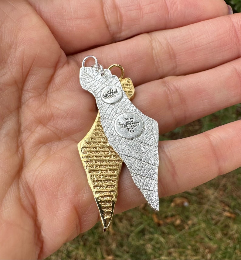 May include: Two pendant necklaces in the shape of Israel. One is silver-colored with a leaf-like texture and the other is gold-colored with a textured surface. Both have small, round charms with Hebrew text. The pendants are hanging from a person's hand.