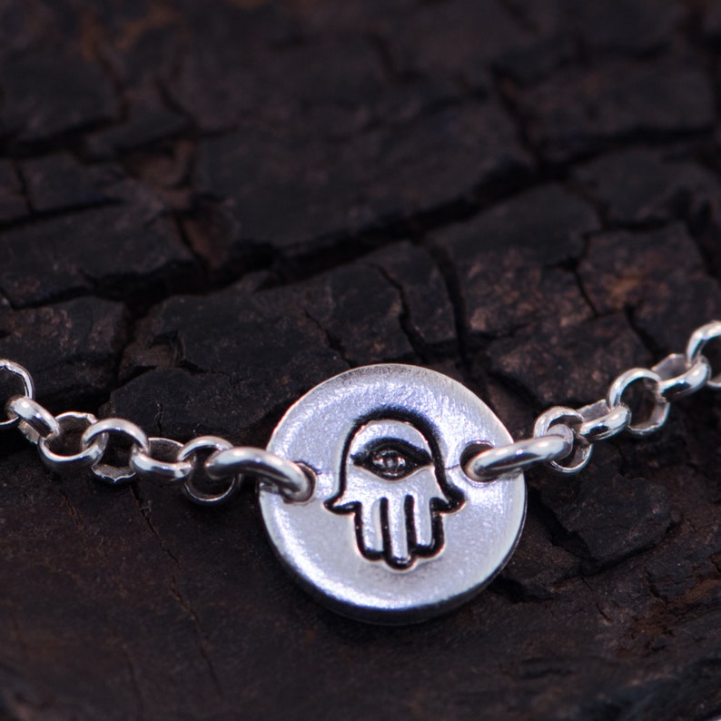 May include: A silver chain bracelet with a round charm featuring a hamsa hand with an eye in the center.