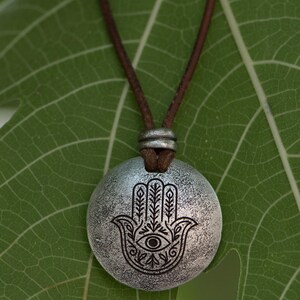 May include: A silver-colored pendant necklace featuring a Hamsa hand design with an eye in the center. The pendant hangs from a brown leather cord. The necklace is displayed on a green leaf.