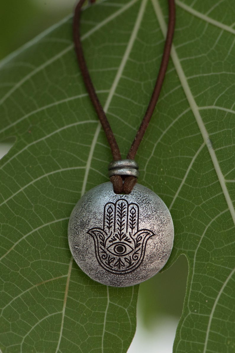 May include: A silver-colored pendant necklace featuring a Hamsa hand design with an eye in the center. The pendant hangs from a brown leather cord. The necklace is displayed on a green leaf.