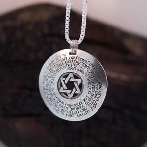 Puede incluir: Un collar con colgante de plata con un colgante circular con texto hebreo y una Estrella de David en el centro. El colgante cuelga de una delicada cadena de plata. El collar es un art&iacute;culo religioso o espiritual.