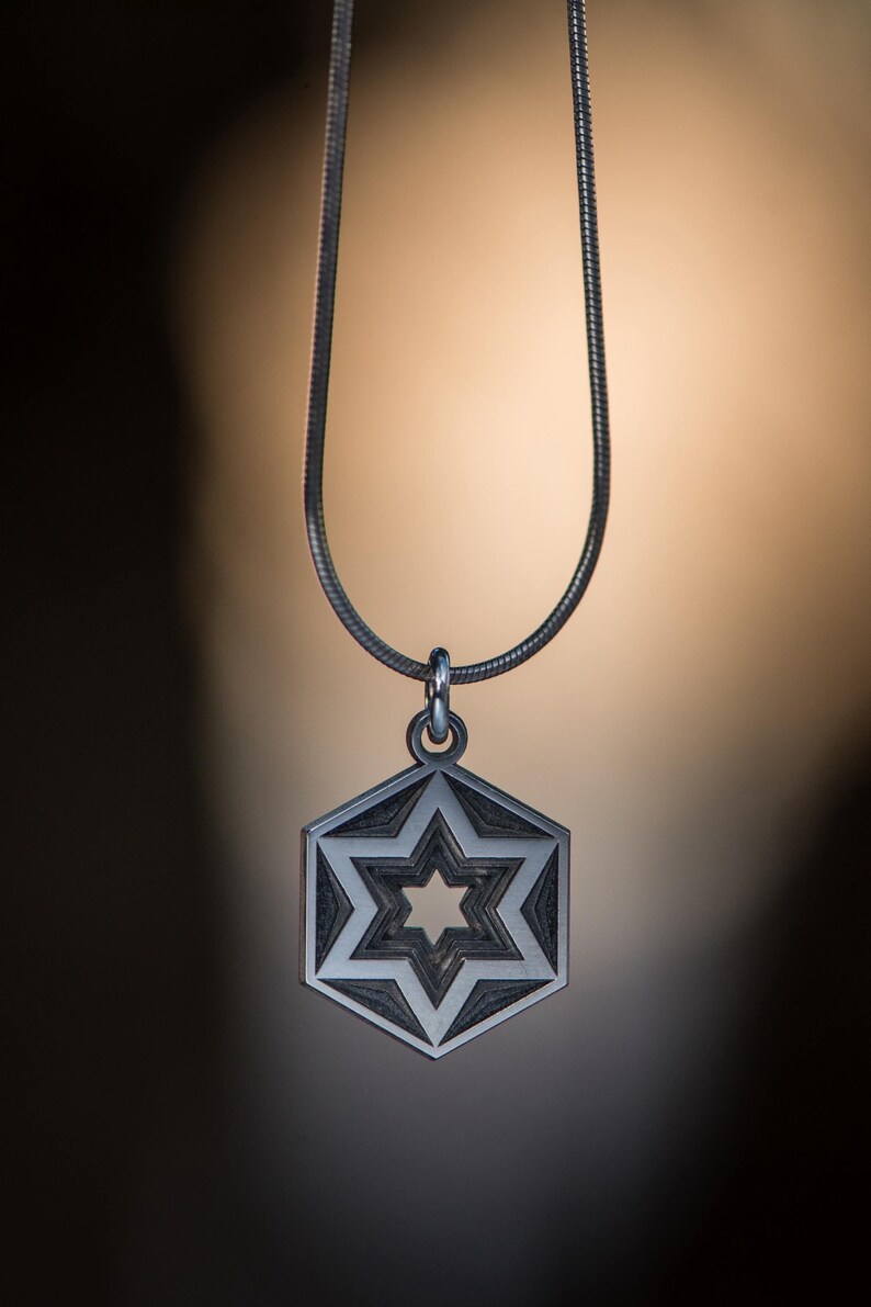 Peut inclure: Un collier argent&eacute; avec un pendentif hexagonal orn&eacute; d'un motif d'&eacute;toile &agrave; six branches. L'&eacute;toile est superpos&eacute;e &agrave; l'int&eacute;rieur de l'hexagone, cr&eacute;ant un motif g&eacute;om&eacute;trique d&eacute;taill&eacute;. La cha&icirc;ne est fine et argent&eacute;e. Le fond est un d&eacute;grad&eacute; de marron et de noir.