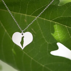 Op de afbeelding: Een zilveren ketting met een hartvormige hanger met een uitgesneden kaart. De ketting hangt tegen een achtergrond van een groen blad, wat een natuurlijke en elegante uitstraling cre&euml;ert. Het ontwerp is eenvoudig en elegant.