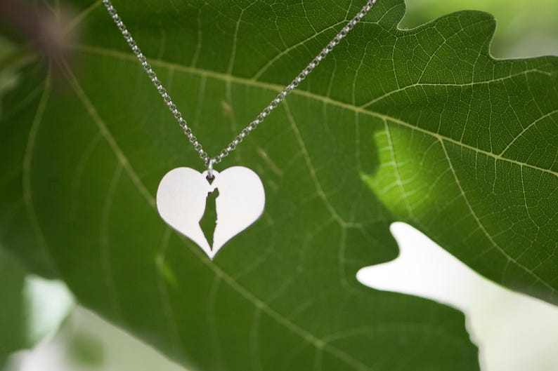 Op de afbeelding: Een zilveren ketting met een hartvormige hanger met een uitgesneden kaart. De ketting hangt tegen een achtergrond van een groen blad, wat een natuurlijke en elegante uitstraling cre&euml;ert. Het ontwerp is eenvoudig en elegant.