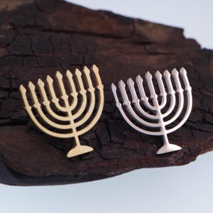 K&ouml;nnte beinhalten: Zwei metallene Menora-Ornamente, eines goldfarben und eines silberfarben, werden auf einer dunklen, strukturierten Oberfl&auml;che gezeigt. Jede Menora hat neun Kerzenhalter und einen zentralen Stiel. Die goldene Menora befindet sich links, die silberne rechts.