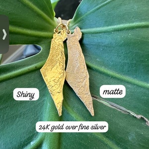 May include: Two gold pendants shaped like the map of Israel, crafted from 24K gold over fine silver. One pendant features a shiny, reflective finish, while the other has a textured, matte finish. The text "Shiny", "matte", and "24K gold over fine silver" is visible.