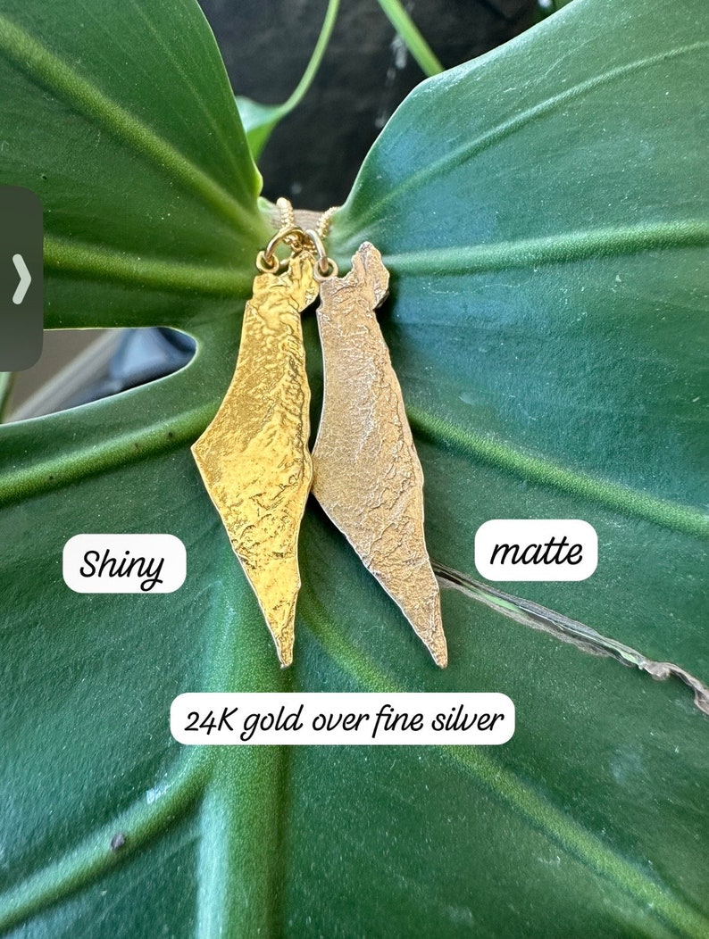 May include: Two gold pendants shaped like the map of Israel, crafted from 24K gold over fine silver. One pendant features a shiny, reflective finish, while the other has a textured, matte finish. The text "Shiny", "matte", and "24K gold over fine silver" is visible.