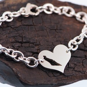 May include: A silver chain link bracelet featuring a heart-shaped charm. The charm has a cutout silhouette of a geographic landmass in the centre. The bracelet is secured with a lobster claw clasp and has a polished, reflective metallic finish.