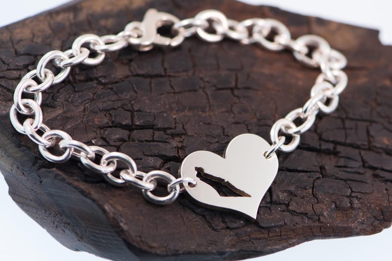 May include: A silver chain link bracelet featuring a heart-shaped charm. The charm has a cutout silhouette of a geographic landmass in the centre. The bracelet is secured with a lobster claw clasp and has a polished, reflective metallic finish.