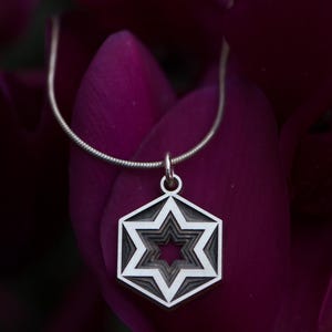 Peut inclure: Un collier avec un pendentif argent&eacute; repr&eacute;sentant une &eacute;toile &agrave; plusieurs couches dans un cadre hexagonal. Le pendentif est suspendu &agrave; une fine cha&icirc;ne argent&eacute;e. Le fond est un violet profond flou, cr&eacute;ant un contraste saisissant.