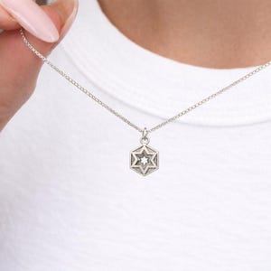 Peut inclure: Un collier argent&eacute; avec un pendentif hexagonal orn&eacute; d'une &eacute;toile. Le pendentif est suspendu &agrave; une fine cha&icirc;ne. Le collier est pr&eacute;sent&eacute; sur un fond blanc.