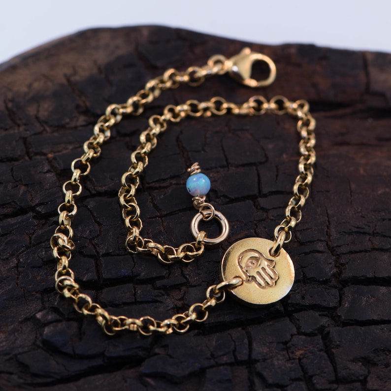 May include: A gold chain bracelet with a hamsa hand charm and a small opal bead. The bracelet is on a dark wood background.