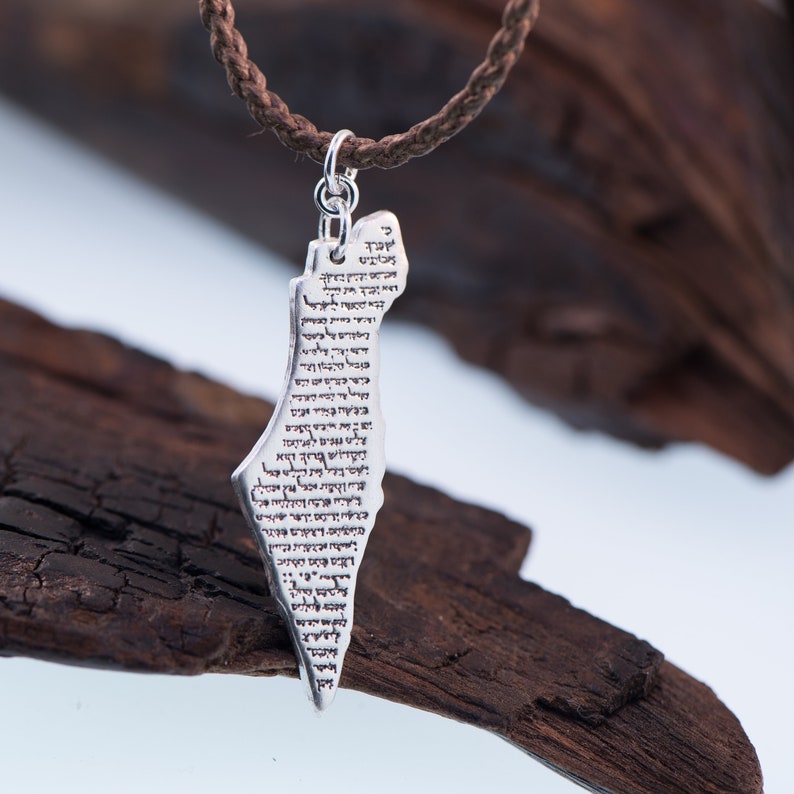 May include: Silver pendant necklace featuring the shape of a country, with text etched on the surface. The pendant hangs from a brown braided cord. The background includes dark brown wood pieces.