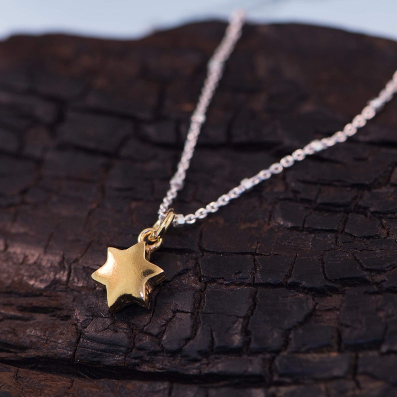 May include: A gold star pendant necklace with a silver chain. The star is a six-pointed shape, and the chain is delicate. The necklace is displayed on a dark, textured surface.