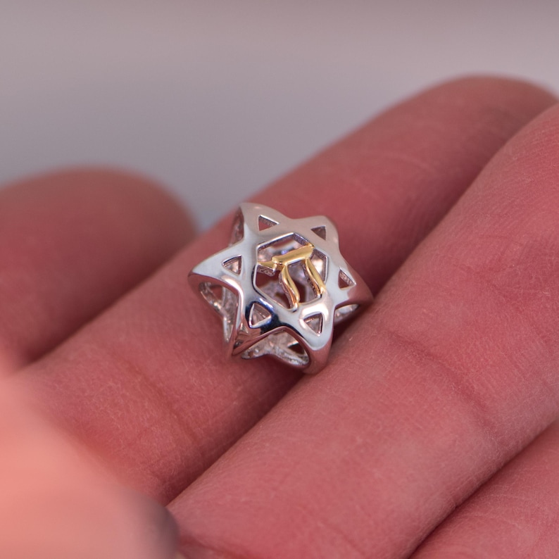 K&ouml;nnte beinhalten: Ein silberner Davidstern-Anh&auml;nger mit einem goldenen hebr&auml;ischen Buchstaben im Inneren. Der Anh&auml;nger befindet sich auf einem Finger.