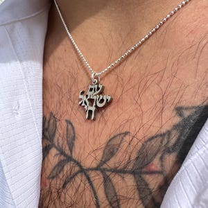 Puede incluir: Un collar de cadena de plata con un colgante con letras hebreas. El colgante tiene forma de &aacute;rbol con ramas y hojas.