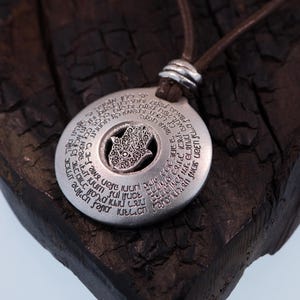 May include: A silver pendant necklace with a brown leather cord. The pendant is circular with engraved text and a central Hamsa hand design. The necklace is displayed on a dark, textured wooden surface.