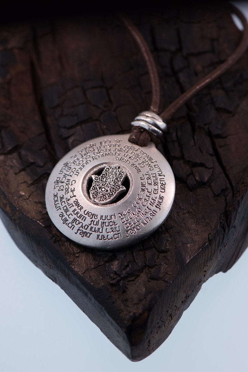 May include: A silver pendant necklace with a brown leather cord. The pendant is circular with engraved text and a central Hamsa hand design. The necklace is displayed on a dark, textured wooden surface.