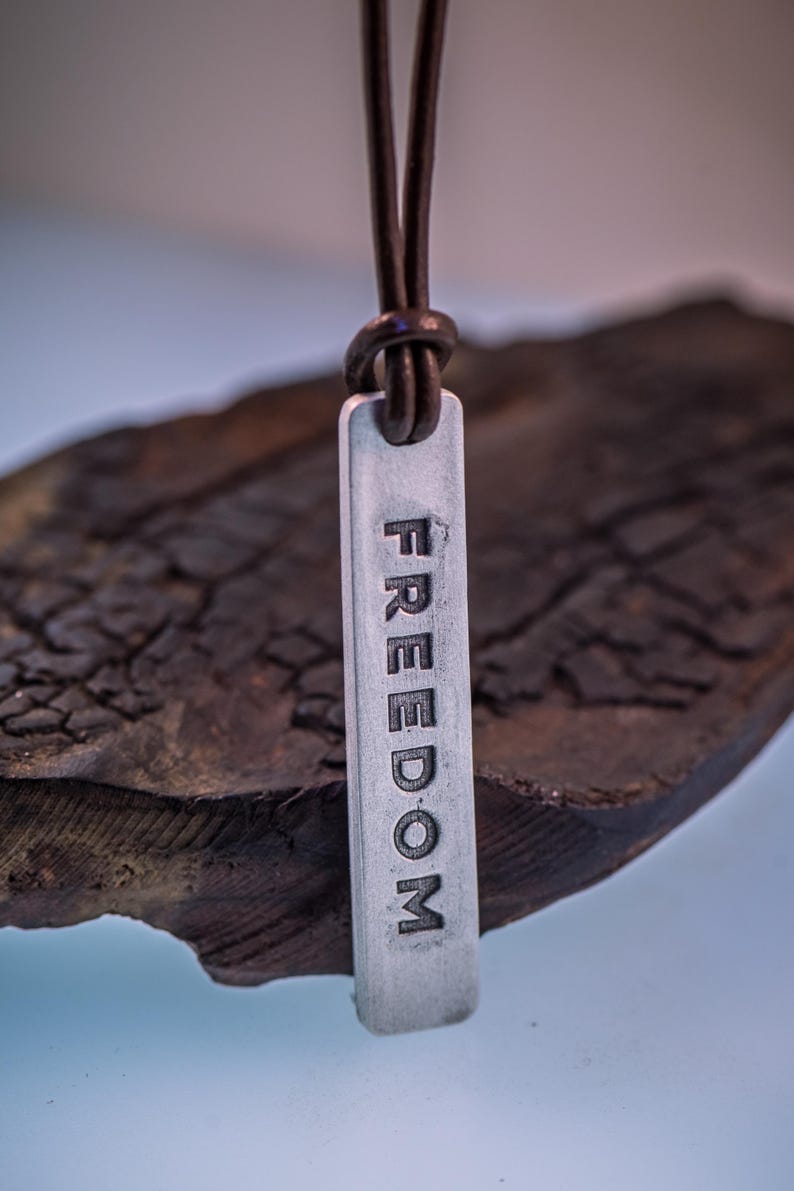 May include: A silver pendant necklace with the word "FREEDOM" engraved vertically. The pendant hangs from a brown leather cord. The background is a dark, textured wooden surface.
