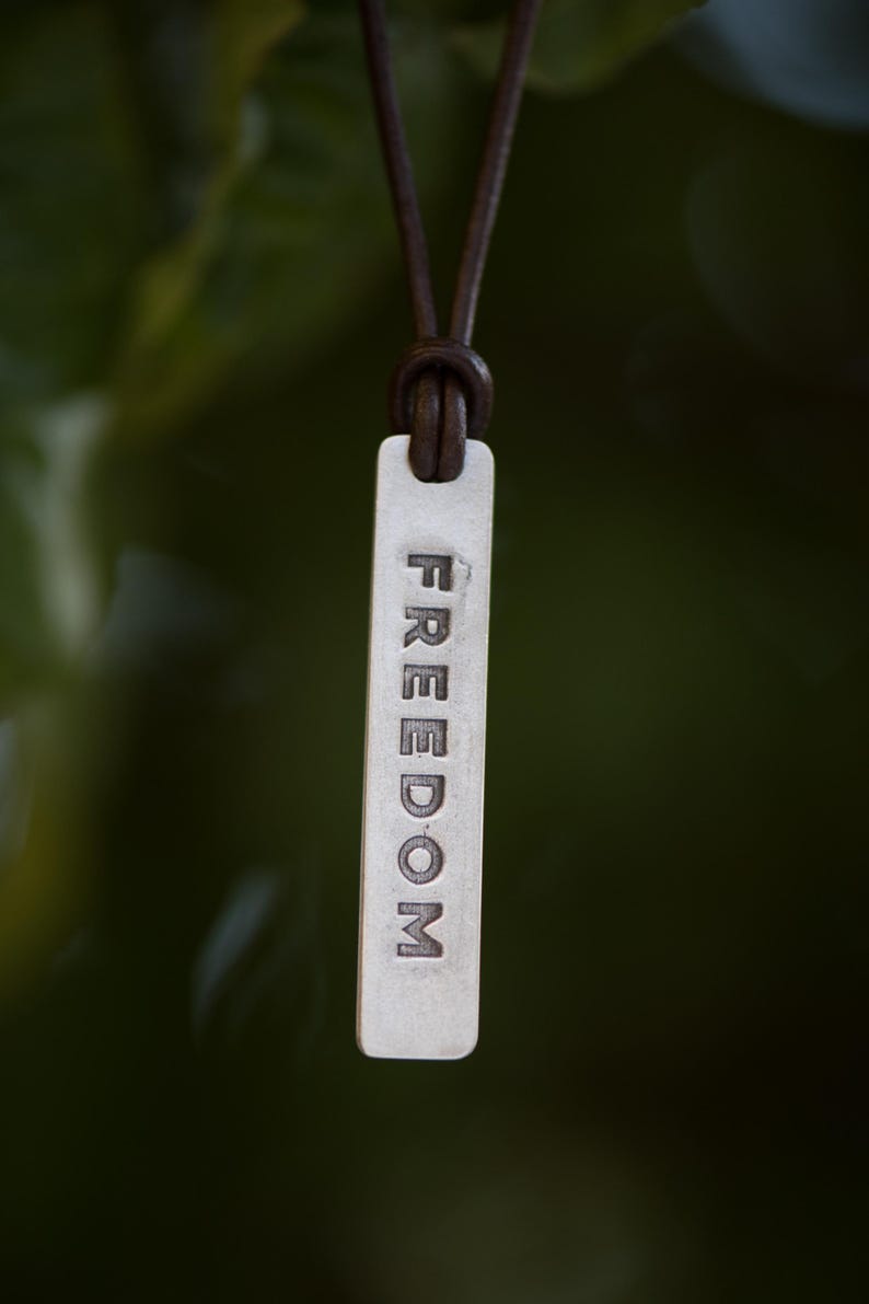 May include: A silver rectangular pendant with the word "FREEDOM" engraved vertically. The pendant is suspended from a brown leather cord. The background is a blurred green.