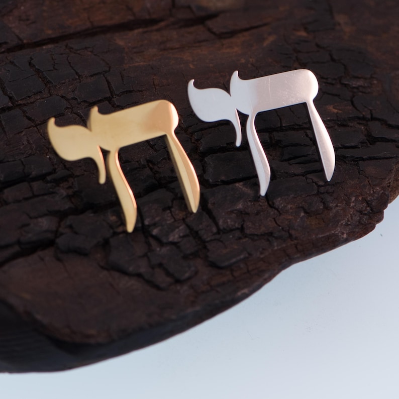 K&ouml;nnte beinhalten: Zwei Chai-Anh&auml;nger aus Metall, einer goldfarben und einer silberfarben, liegen auf einer dunklen, strukturierten Holzoberfl&auml;che. Die Anh&auml;nger haben die Form des hebr&auml;ischen Buchstabens Chai, der das Leben symbolisiert. Der goldfarbene Anh&auml;nger befindet sich links, der silberfarbene rechts.