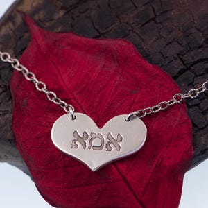 May include: A silver heart-shaped pendant necklace with the Hebrew word "אמא" engraved on it. The pendant hangs from a delicate chain. The necklace rests on a dark red leaf against a dark wooden background.