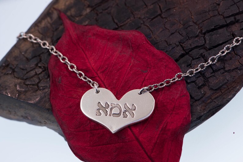 May include: A silver heart-shaped pendant necklace with the Hebrew word "אמא" engraved on it. The pendant hangs from a delicate chain. The necklace rests on a dark red leaf against a dark wooden background.