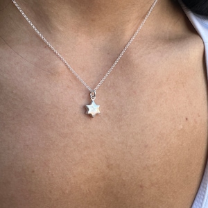 May include: A silver necklace featuring a small, six-pointed star pendant. The star has a textured finish and is suspended from a fine chain. The necklace is worn against the skin, with a white top visible.