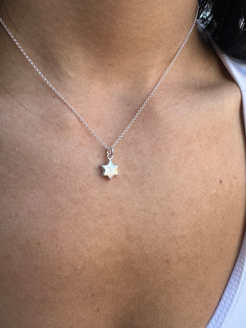 May include: A silver necklace featuring a small, six-pointed star pendant. The star has a textured finish and is suspended from a fine chain. The necklace is worn against the skin, with a white top visible.