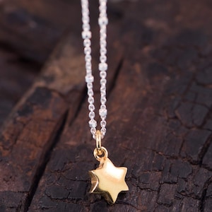 May include: A silver chain necklace with a gold-coloured star pendant. The star has a textured surface and is suspended from a small loop. The necklace is set against a dark, textured background.