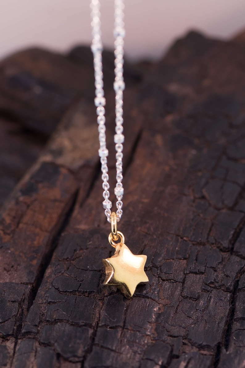 May include: A silver chain necklace with a gold-coloured star pendant. The star has a textured surface and is suspended from a small loop. The necklace is set against a dark, textured background.