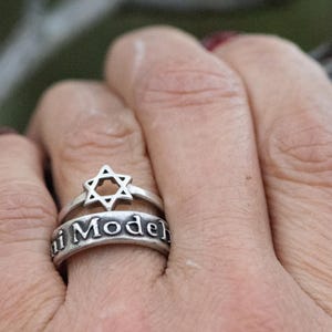 Puede incluir: Un conjunto de anillos de plata con un dise&ntilde;o de Estrella de David y la inscripci&oacute;n "i Model". El anillo se lleva en un dedo, mostrando los intrincados detalles de la joya. El anillo es un s&iacute;mbolo de fe y herencia.