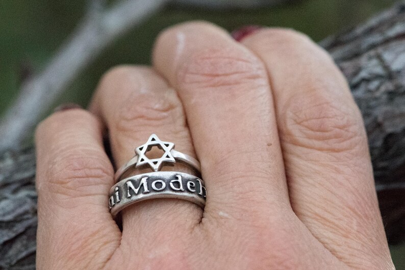 Puede incluir: Un conjunto de anillos de plata con un dise&ntilde;o de Estrella de David y la inscripci&oacute;n "i Model". El anillo se lleva en un dedo, mostrando los intrincados detalles de la joya. El anillo es un s&iacute;mbolo de fe y herencia.
