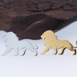 May include: Two lion-shaped metal pins. One is silver-colored, the other is gold-colored. Both pins are side-profile depictions of a lion with a flowing mane and a curled tail. The pins are on a white surface with a dark brown, textured background.
